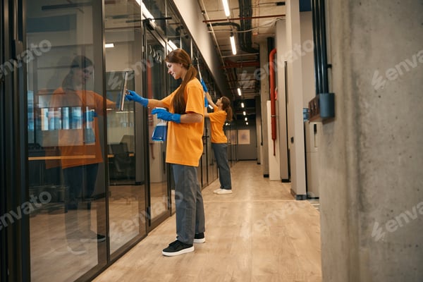 Zwei Beschäftigte in Arbeitskleidung mit Reinigungsutensilien in einem modernen Büroflur