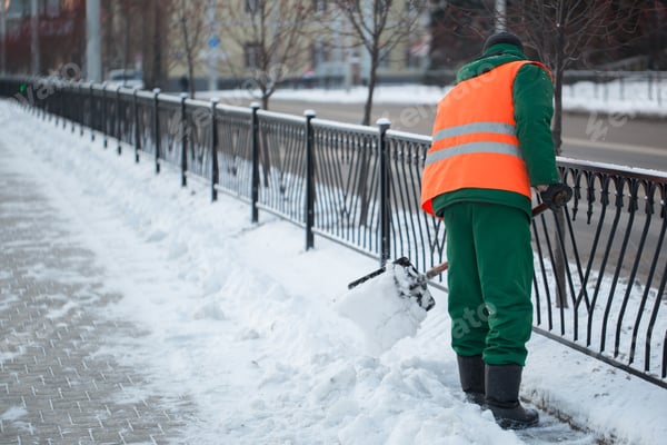 Person räumt Schnee auf einem Gehweg entlang eines Zauns